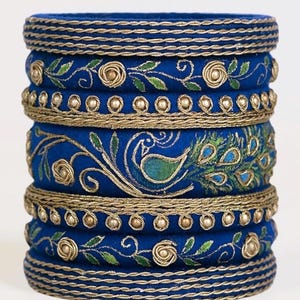 May include: A stack of blue fabric bangles with gold and green embroidery. The bangles feature floral and peacock designs, with gold trim and small, round embellishments. The overall aesthetic is ornate and decorative.