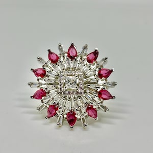 Peut inclure: Une bague argentée avec un carré central de pierres claires, entouré d'un carré de petites pierres roses. La bague présente des pierres rectangulaires claires et des pierres en forme de goutte rouge rubis alternées, créant un motif en étoile.