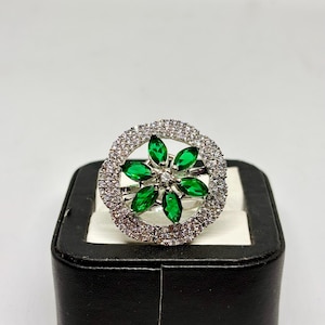 Peut inclure: Bague en argent ornée d'un motif floral avec des pétales en pierres vertes et une pierre centrale transparente. Les pétales sont entourés d'un halo de petites pierres transparentes. La bague est présentée sur un écrin noir.