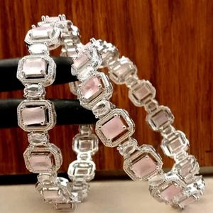 以下が含まれることがあります： 銀色のバングル2本。質感のある装飾的な金属フレームにセットされた長方形の淡いピンク色の石が繰り返されるパターンが特徴です。各石の両脇には、より小さな透明な楕円形の石が配置され、連結したデザインを作り出しています。