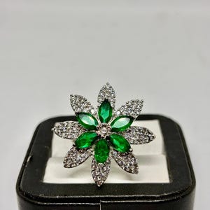Peut inclure: Bague argentée en forme de fleur, ornée de pierres précieuses vertes et blanches. Le centre de la fleur est orné d'un petit diamant clair. La bague est présentée sur un écrin noir. Les pierres sont disposées en motif floral.