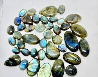 May include: A selection of polished labradorite stones in diverse shapes and sizes. The stones exhibit iridescent flashes of blue, green, and gold, with oval, teardrop, and irregular forms. The stones are arranged on a white surface.