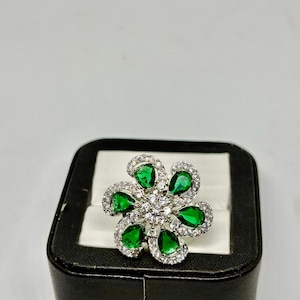Peut inclure: Une bague argentée avec un motif floral. Les pétales de la bague sont formés de pierres vertes en forme de goutte, chacune entourée de petites pierres claires. Le centre de la fleur est rempli de pierres claires supplémentaires. La bague est présentée sur un écrin noir.