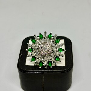 Peut inclure: Bague argentée ornée d'une pierre centrale carrée transparente, entourée de pierres transparentes taillées en baguette et de pierres vertes en forme de goutte. La bague est présentée sur un écrin noir à intérieur blanc.