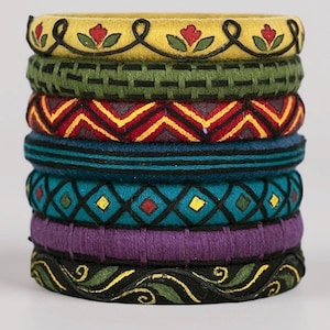 May include: A stack of seven colorful fabric bangles. The bangles feature various patterns and colors, including yellow with red floral accents, green crosshatch, red and yellow zigzags, teal, turquoise diamond patterns, purple, and black with green swirls.