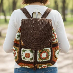 Op de afbeelding: Een bruine gehaakte rugzak met een klepsluiting en verstelbare bandjes. De tas heeft een patchwork-ontwerp met vierkanten in bruin, oranje, groen en wit. De rugzak is gemaakt van garen en heeft een trekkoordsluiting.