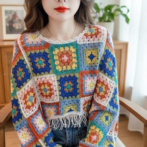 Op de afbeelding: Een kleurrijke gehaakte top met een patchwork-ontwerp met vierkanten in tinten blauw, oranje, geel en roze. De top heeft een ronde halslijn met witte kanten bies en een gefranjerde zoom. De mouwen zijn lang.