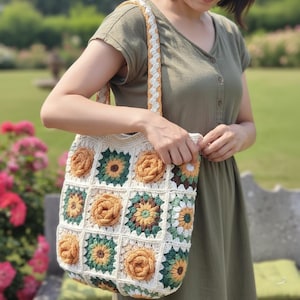 May include: A cream-colored crochet tote bag with a sunflower design. The bag features square panels with orange, green, and yellow floral patterns. The strap is braided with cream, orange, and yellow yarn. The bag is held by a person.