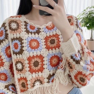Op de afbeelding: Een kleurrijke gehaakte top met een vierkant patroon en bloemmotieven in oranje, bruin, blauw en crème. De top heeft lange mouwen met franjes aan de zoom en manchetten. De halslijn is geschulpt.