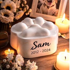 Op de afbeelding: Een witte urn voor huisdieren in de vorm van een pootafdruk, met de naam "Sam" en de data 2012-2024. De afbeelding toont een ingelijste foto van een hond, kaarsen en witte bloemen, wat een gevoel van herinnering oproept.