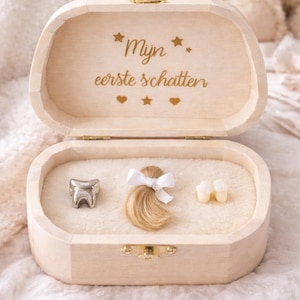 Op de afbeelding: Een houten bewaardoos met de tekst "Mijn eerste schatten" in gouden letters. Binnenin liggen een zilveren tand, een lok blond haar met een witte strik en twee melktanden op een zacht, crèmekleurig oppervlak.