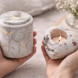 Op de afbeelding: Een urn met marmerpatroon en een hartvormige kandelaar. De urn is cilindrisch met een deksel en de kandelaar heeft een brandende kaars. Beide items hebben gouden accenten. De items worden in handen gehouden.