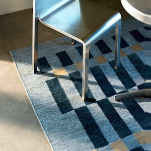 May include: A modern, geometric rug in shades of blue, navy, and tan. The rug features a repeating pattern of rectangular and square shapes. A silver metal chair and a small table are partially visible in the image, suggesting a contemporary interior design.