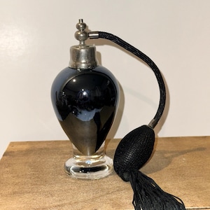 May include: An antique perfume bottle with a black, pear-shaped body and a silver-toned metal top. The bottle has a clear glass base and a black atomizer with a tassel. The bottle is displayed on a wooden surface.