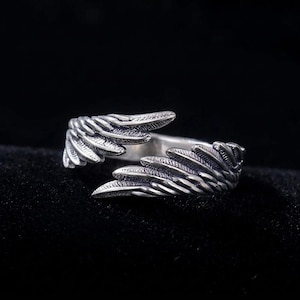 Op de afbeelding: Een zilverkleurige ring met een vleugelontwerp. De ring heeft twee gedetailleerde vleugels die zich om de vinger wikkelen. De vleugels hebben getextureerde veerd details, waardoor een realistische uitstraling ontstaat. De ring staat tegen een donkere achtergrond.