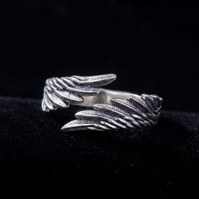 Op de afbeelding: Een zilverkleurige ring met een vleugelontwerp. De ring heeft twee gedetailleerde vleugels die zich om de vinger wikkelen. De vleugels hebben getextureerde veerd details, waardoor een realistische uitstraling ontstaat. De ring staat tegen een donkere achtergrond.