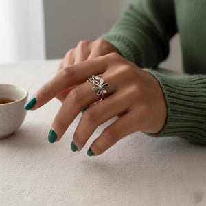 May include: A silver ring with a double daisy flower design. The ring is worn on a hand with green nail polish, set against a green jumper and a white surface. The floral ring design adds a touch of nature-inspired elegance.