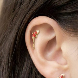 May include: Gold-coloured koi fish earrings with red and white details. The earrings, shaped like the fish, are worn on the ear. A unique and stylish accessory, these earrings are a great addition to any jewellery collection.