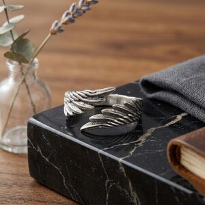 Op de afbeelding: Een zilverkleurige ring met een vleugelontwerp. De open ring wikkelt zich om de vinger, met gedetailleerde veertjes. Hij ligt op een zwart marmeren oppervlak, naast een kleine glazen vaas met een lavendel takje.