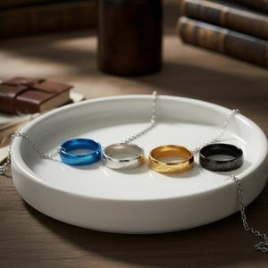 May include: Four rings on silver chains, presented on a white ceramic plate. The rings are blue, silver, gold, and black. The gold ring has engraved text. The background includes books and a small leather-bound book.