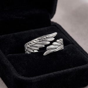 Op de afbeelding: Een zilverkleurige ring in de vorm van engelen vleugels, gepresenteerd in een zwarte sieradendoos. De ring heeft gedetailleerde veertjesstructuren en een open ontwerp voor verstelbare maat. Het is een sieraad.