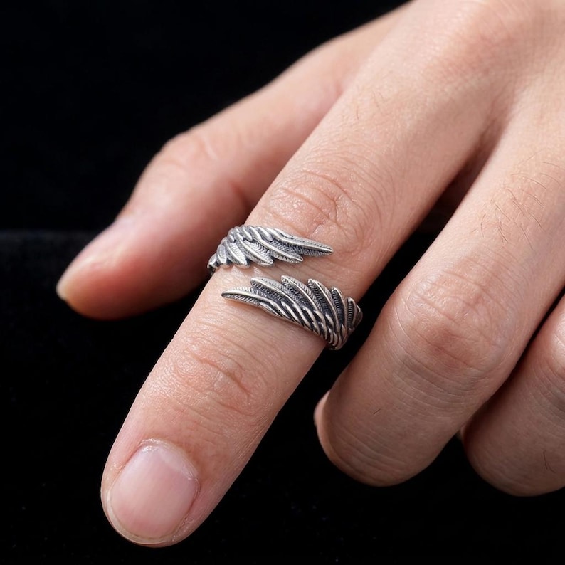 Op de afbeelding: Een zilverkleurige ring met een vleugelontwerp. De ring wikkelt zich om de vinger, waarbij elke vleugelpunt naar buiten uitsteekt. De veren van de ring zijn gedetailleerd en het metaal heeft een licht verouderde uitstraling. De ring is een sieraad.