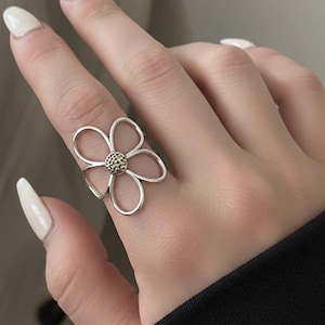 May include: A silver-colored flower ring with a textured center, worn on a finger. The ring's petals are open and delicate. The text "Design by Sajeel" is visible at the bottom.