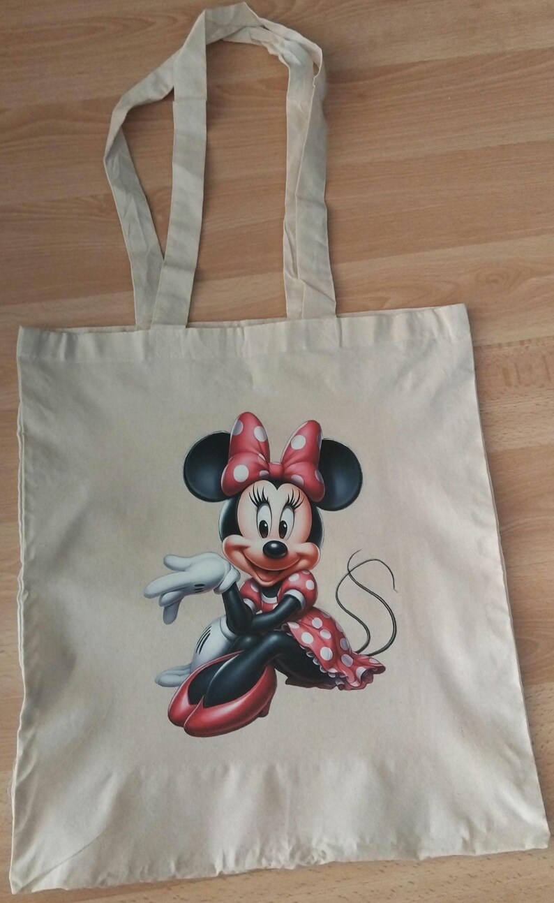 Puede incluir: Un bolso tote de lona beige con una colorida ilustraci&oacute;n de Minnie Mouse. Minnie est&aacute; representada con un vestido y lazo de lunares rojos, con guantes blancos y zapatos rojos. El bolso tiene dos asas beige.