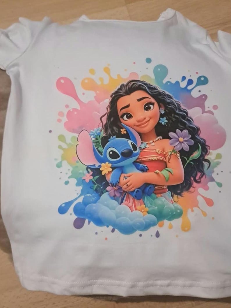 May include: White t-shirt featuring a colorful graphic of Moana and Stitch. Moana is depicted with long black hair, wearing a red top and holding a blue Stitch plush. The background includes colorful paint splatters and floral accents.