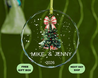 May include: A clear, faceted glass Christmas ornament with a green ribbon. The ornament features a watercolor painting of a Christmas tree with a red and white bow. The names "MIKE & JENNY" and the year "2026" are printed on the ornament.