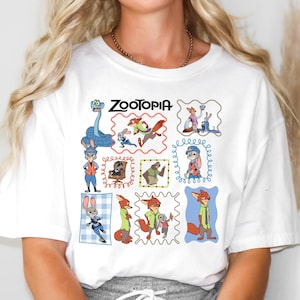 May include: White t-shirt featuring the word "ZOOTOPIA" above a grid of colorful cartoon character illustrations. The illustrations include a blue snake, a rabbit, and a fox. The shirt is made of a soft material.