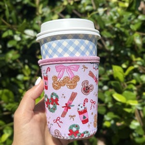 Peut inclure: Une pochette à café en néoprène violet clair avec un motif répétitif d'icônes sur le thème des fêtes, incluant des biscuits en pain d'épices, des sucres d'orge, des couronnes et des nœuds roses, entourant un gobelet avec un couvercle blanc.