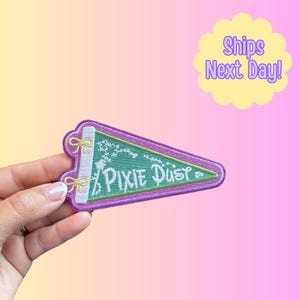 May include: A purple and green pennant-shaped patch with the words "Pixie Dust" in white script. The patch has a white border with gold bows and a green background with white decorative elements. The background is a gradient of yellow, pink, and purple.