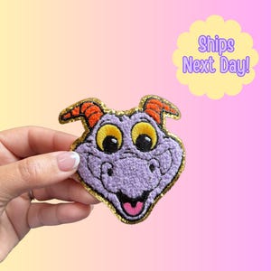 May include: Embroidered patch featuring a purple dragon-like character with large yellow eyes, orange horns, and a gold glitter border. The character has a smiling expression. The patch is held against a gradient background.