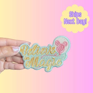 May include: Embroidered patch with the words "Believe in Magic" in gold script on a light blue background. A pink Mickey Mouse balloon design is on the right. The patch has a light blue border.
