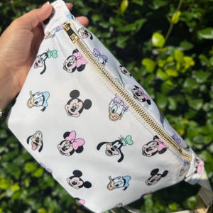 May include: A white fanny pack featuring a repeating pattern of Disney character faces including Minnie Mouse, Mickey Mouse, Donald Duck, Daisy Duck, and Goofy. The bag has a gold-toned metal zipper closure and an adjustable white fabric strap.