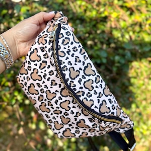 K&ouml;nnte beinhalten: G&uuml;rteltasche mit Leopardenmuster mit hellbraunen und schwarzen Flecken auf wei&szlig;em Grund. Die Tasche hat einen schwarzen Riemen und einen goldenen Rei&szlig;verschluss. Ein modisches Accessoire.