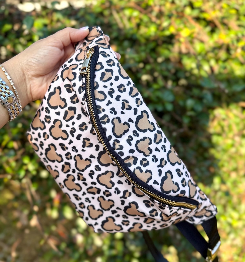K&ouml;nnte beinhalten: G&uuml;rteltasche mit Leopardenmuster mit hellbraunen und schwarzen Flecken auf wei&szlig;em Grund. Die Tasche hat einen schwarzen Riemen und einen goldenen Rei&szlig;verschluss. Ein modisches Accessoire.