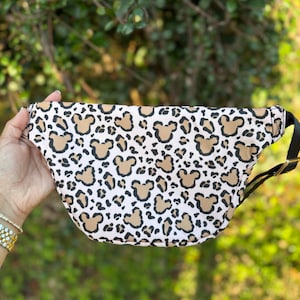 K&ouml;nnte beinhalten: G&uuml;rteltasche mit einem Muster aus braunen Mickey Mouse-K&ouml;pfen und Leopardenflecken auf wei&szlig;em Grund. Die Tasche hat einen schwarzen Riemen und eine gebogene Form. Die Tasche wird von einer Person gehalten.