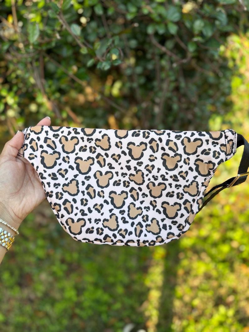 K&ouml;nnte beinhalten: G&uuml;rteltasche mit einem Muster aus braunen Mickey Mouse-K&ouml;pfen und Leopardenflecken auf wei&szlig;em Grund. Die Tasche hat einen schwarzen Riemen und eine gebogene Form. Die Tasche wird von einer Person gehalten.