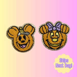 May include: Two embroidered patches featuring orange pumpkin-shaped characters with mouse-like ears and gold glitter borders. The left patch shows a character with a small hat, while the right patch features a character wearing a purple bow.