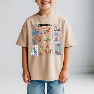 May include: Tan t-shirt featuring the word "ZOOTOPIA" above a grid of cartoon character illustrations. The shirt has short sleeves and is worn by a child. The illustrations include a snake, a bunny, and a fox.