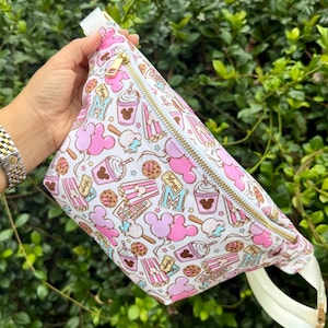 May include: A white fanny pack featuring a repeating pattern of pink Mickey Mouse-shaped balloons, popcorn buckets, cookies, and drinks with the text Magic. The bag has a gold-tone zipper closure and an adjustable white strap with a plastic buckle.
