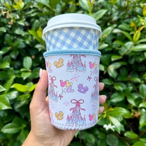 May include: A white fabric cup sleeve featuring a repeating pattern of light blue castles, pink bows, and colorful Mickey Mouse-shaped balloons. The sleeve has a light blue trim and is placed around a cup with a light blue and white gingham lid.