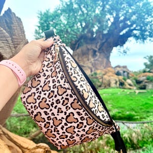 K&ouml;nnte beinhalten: Eine rosa-beige Bauchtasche mit einem sich wiederholenden Mickey Mouse-Kopfmuster und einem goldenen Rei&szlig;verschluss. Die Tasche hat einen schwarzen Riemen und eine Schnalle. Der Hintergrund zeigt einen gro&szlig;en Baum und gr&uuml;nes Laub.
