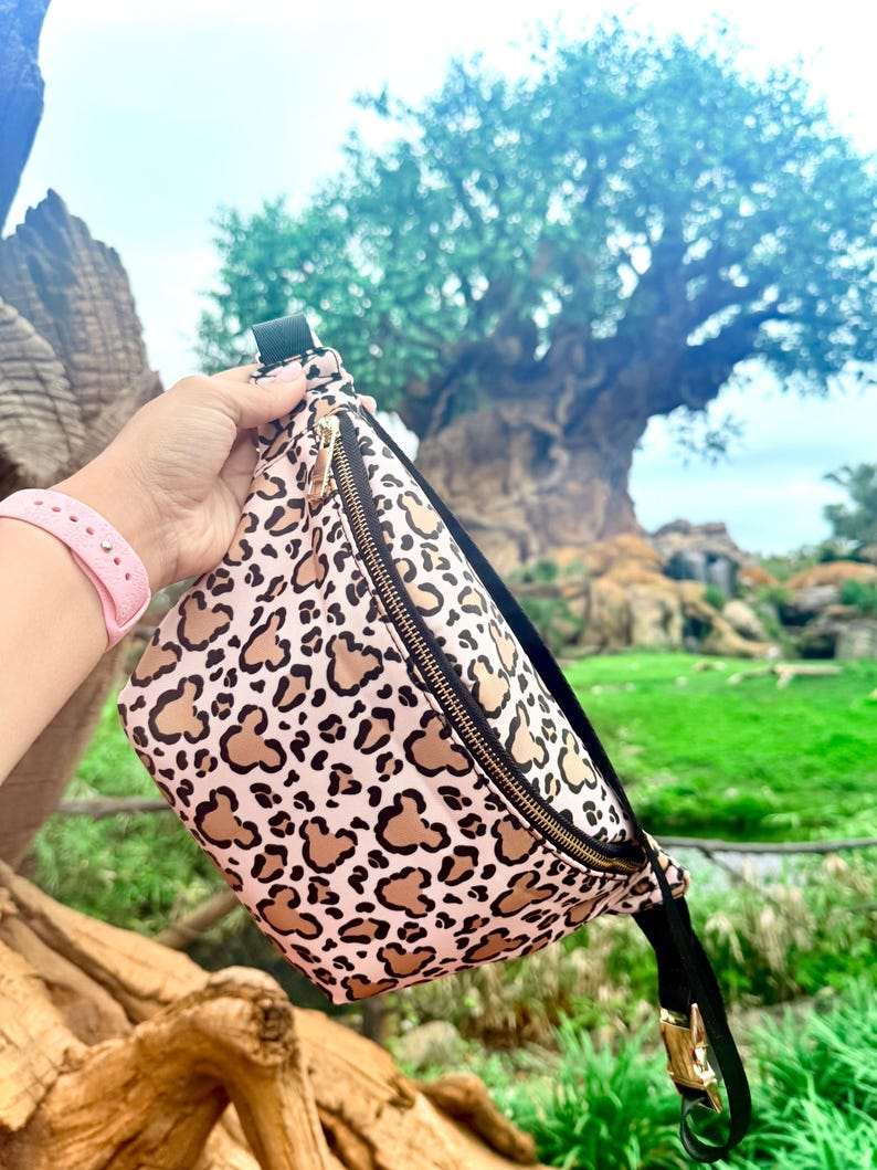 K&ouml;nnte beinhalten: Eine rosa-beige Bauchtasche mit einem sich wiederholenden Mickey Mouse-Kopfmuster und einem goldenen Rei&szlig;verschluss. Die Tasche hat einen schwarzen Riemen und eine Schnalle. Der Hintergrund zeigt einen gro&szlig;en Baum und gr&uuml;nes Laub.