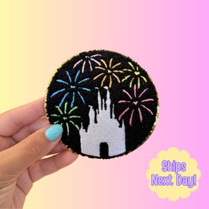 May include: A circular embroidered patch featuring a white castle silhouette against a black background, surrounded by colorful fireworks in shades of blue, green, yellow, and pink. The patch has a gold-colored border and a textured fabric finish.