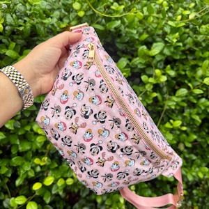May include: A light pink fanny pack featuring a repeating pattern of Disney characters including Mickey Mouse, Minnie Mouse, Donald Duck, and Pluto wearing winter hats and scarves, finished with a gold-toned metal zipper and a matching pink strap.