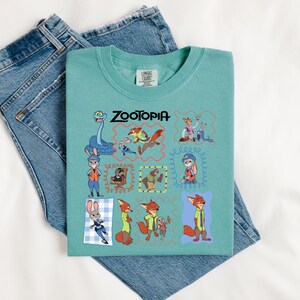 May include: A teal Comfort Colors t-shirt featuring the word "ZOOTOPIA" in black text. The shirt has a colorful graphic of characters from the animated movie. The shirt is laid on top of a pair of blue denim jeans.