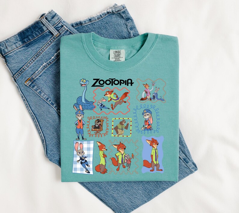 May include: A teal Comfort Colors t-shirt featuring the word "ZOOTOPIA" in black text. The shirt has a colorful graphic of characters from the animated movie. The shirt is laid on top of a pair of blue denim jeans.
